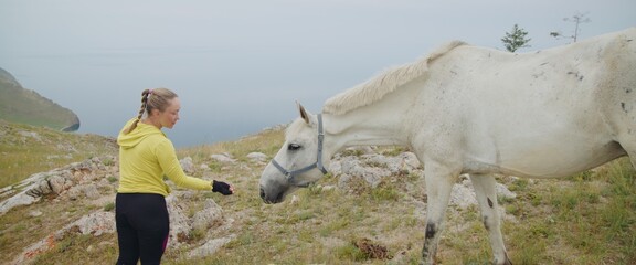 The woman feed domestic white horse on rocky steppe area on top of mountain. The girl looks and...
