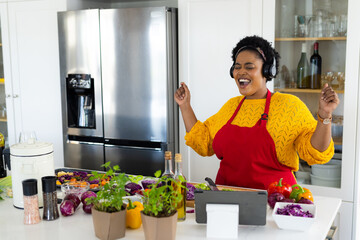 Plus size african american woman in apron preparing meal and singing using tablet in kitchen