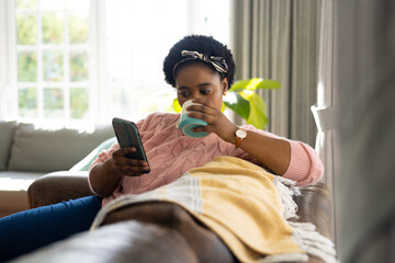 Plus size african american woman drinking coffee and using smartphone in living room
