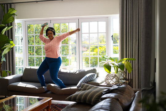 Happy plus size african american woman with headphones and smartphone dancing on sofa