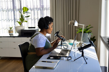 Happy plus size african american woman using headphones, laptop and microphone podcasting from home