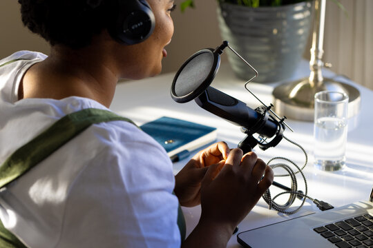 Happy plus size african american woman using headphones, laptop and microphone podcasting from home