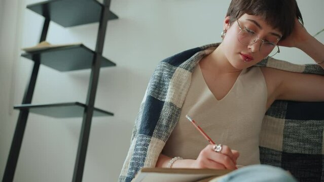 Pensive woman covered with blanket drawing in notebook at home
