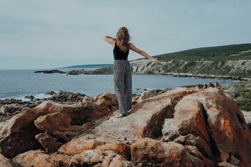 Young female standing on a rocky shoreline, taking in the view of the vast blue expanse of the ocean