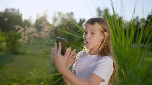 Little smiling cute girl using smartphone outdoors, make video call on mobile. Emotions good news surprise concept. Kid girl holding smart phone enjoying technology device, talking by call on phone.