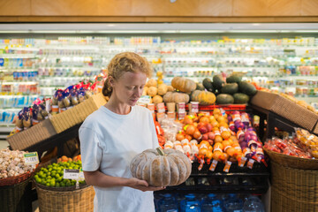 
A young girl of European appearance with blond curly hair holds a pumpkin in her hands while standing at a supermarket counter.