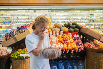 
A young girl of European appearance with blond curly hair holds a pumpkin in her hands while standing at a supermarket counter.