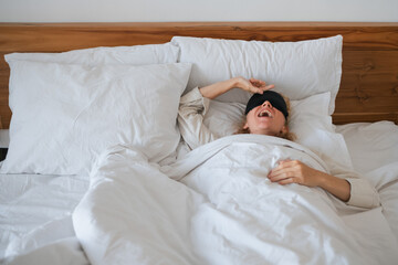 A young girl, of European appearance, with blond, curly hair, wakes up in a sleep mask, on a bed, under a blanket, on a pillow.