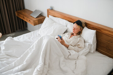 A young girl, of European appearance with blond hair, remotely works on a mobile phone, lying in bed under a blanket on a pillow.