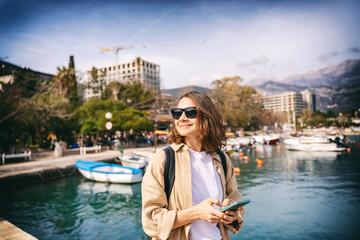 Young stylish happy woman in sunglasses standing on the sea promenade in the city with a smartphone © olezzo