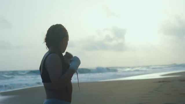 Female boxer MMA fighter girl boxing on beach at sunset sunrise. Back view.