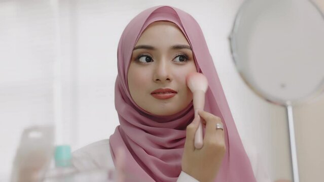 Young asian beautiful muslim woman with hijab looking mirror applying makeup with brusher of cheek daily routine at home. Beautiful of girl holding brush make-up on face.