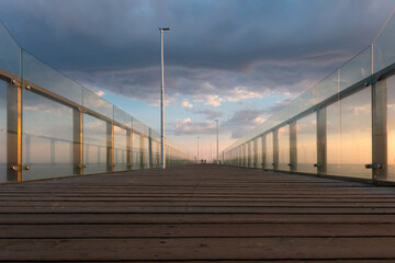 Looking at the perspective of a long pier on the sea in the early morning. There are side protective glass walls and brown plank flooring. Clouds and light from the sunrise are visible in the sky. Bac