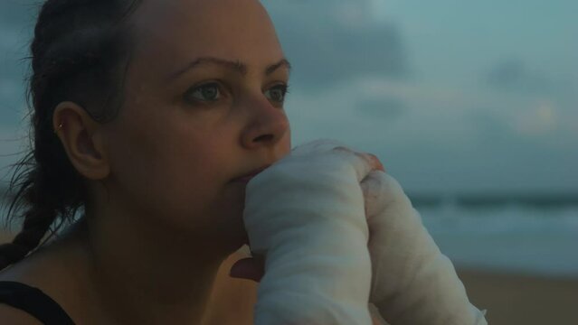 Sad female boxer MMA fighter on beach looks into distance with clasped hands.