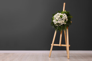Funeral wreath of flowers on wooden stand near dark grey wall indoors, space for text