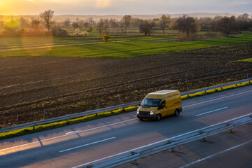 Yellow modern delivery small shipment cargo courier van moving fast on motorway road to city urban suburb. Beautiful sunset in the background.

