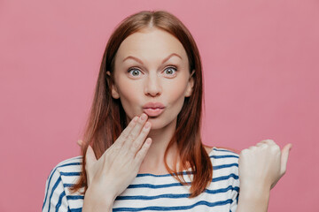 Attractive woman raises eyebrows, pouts lips, hand near mouth. Brown hair. Surprised expression. Pink background.