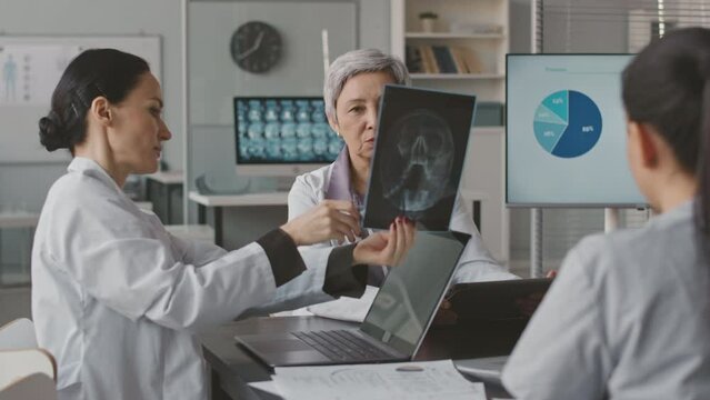 Waist up of multiethnic team of professional doctors analyzing x-ray image of patient sitting together at desk in medical office