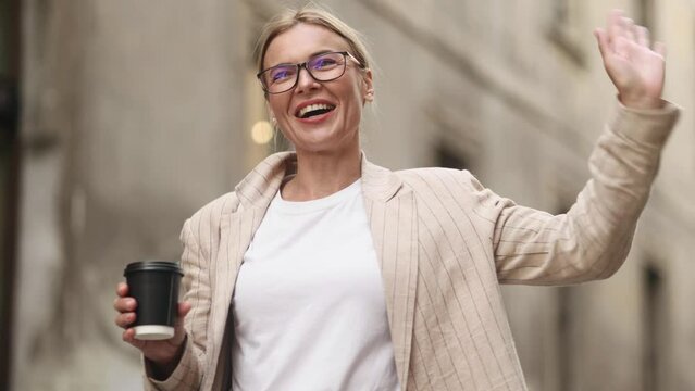 Positive friendly mature blond woman hurry to work with cup of hot coffee or tea in the morning and say hi nodding head greeting wave to colleague at city street Dream job concept