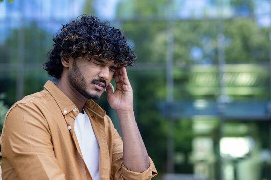 Close-up photo. A young Indian man fell ill on the street outside. He holds his head with his hand, feels severe pain, pressure and stress. He closes his eyes and grimaces from the tension