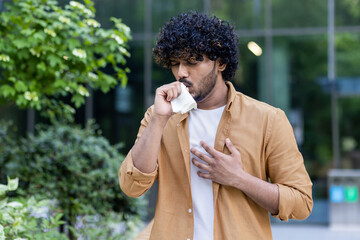 A young Indian man is standing outside on the street and coughing. covers his mouth with a napkin,...