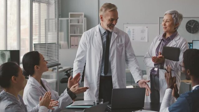 Medium shot of diverse medical team wearing scrubs and lab coats applauding to their adult Caucasian male chief physician while having conference meeting in modern office