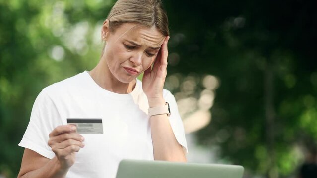 Confused blond woman with credit card thoughtfully pondering on screen while has problem with online order payment error or blocked bank account doing shopping at internet on laptop at park