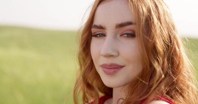 Close up flirty playful woman with glamorous makeup smiles flirtatiously looking at camera. Face of flirtatious glamorous red-haired woman in field in rays of sun. Close up portrait of redhead woman