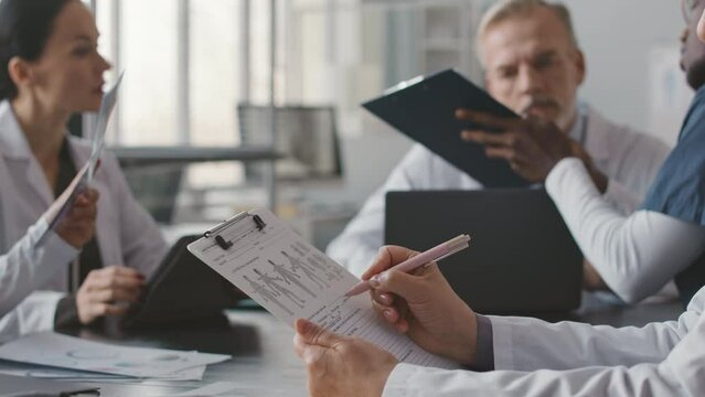Waist up of adult Asian female doctor doing paperwork while having staff meeting with medical team and chief physician in conference room