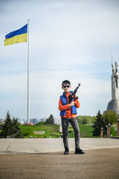 Boy make a picture on monument motherland and Ukrainian flag background. Kid hold in his arms toy machine gun. Camouflage hat and sunglasses