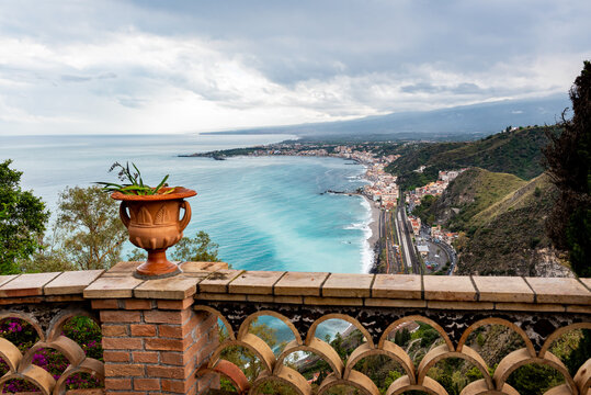 Mediterranean flowering plant in a stone ceramic pot with scenic view on seaside and Mount Etna volcano from public garden Parco Duca di Cesaro to Giardini Naxos in Taormina, Sicily, Italy
