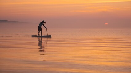 Man paddling on stand up paddle board against background of rising sun. SUP