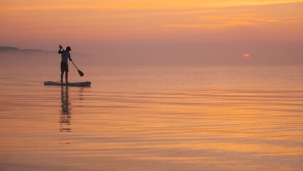 Man paddling on stand up paddle board against background of rising sun. SUP - obrazy, fototapety, plakaty