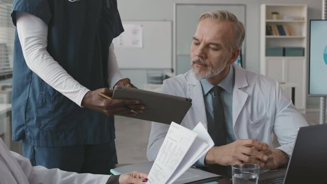 Cropped shot of unrecognizable Black male nurse with digital tablet in hands consulting with adult Caucasian male doctor while having meeting in office