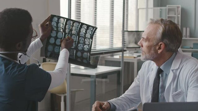 Two diverse male doctors looking at MRI of patient brain and having discussion sitting in modern medical office