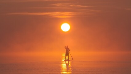 Man paddling on stand up paddle board against background of rising sun. SUP