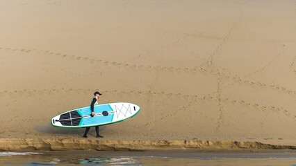 Man walking on shore with stand up paddle board along big dunes in desert. SUP