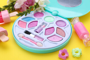 Decorative cosmetics for kids. Eye shadow palette, lipsticks, accessories and flowers on yellow background, closeup