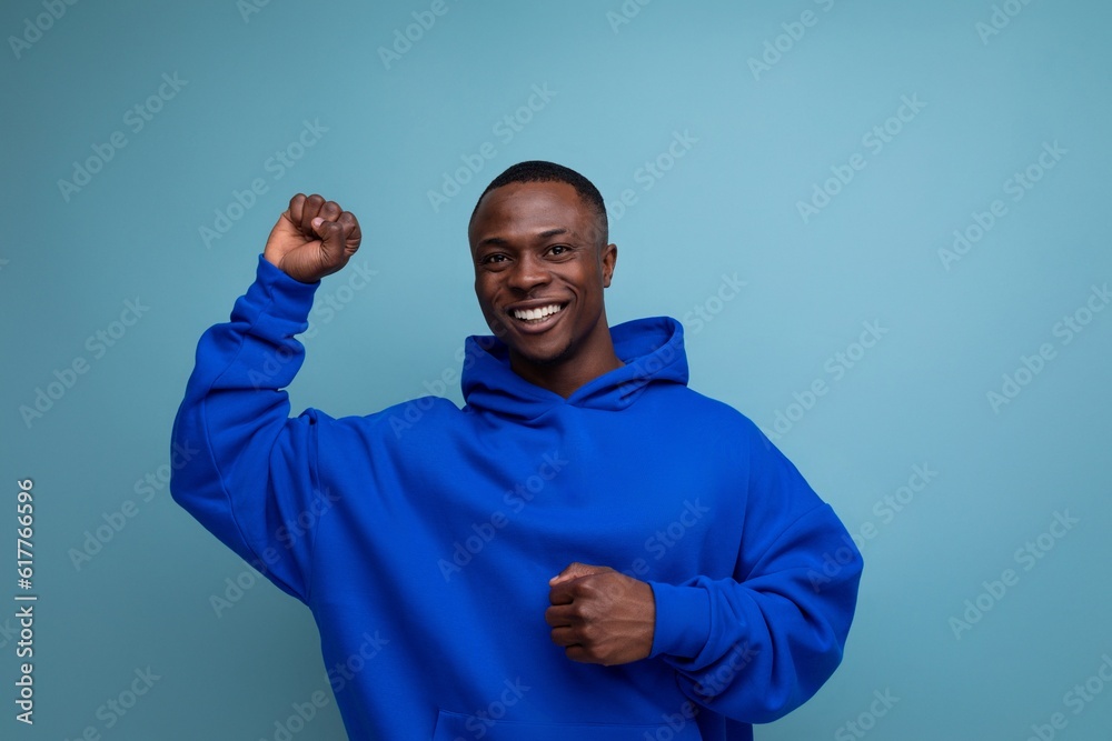 Wall mural cheerful cute ethnic 25 year old african man in a stylish hoodie with a hood on a blue background