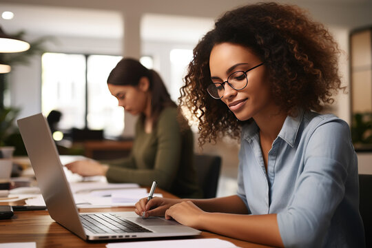 Portrait of Young Biracial Woman Taking Notes at Her Desk While Looking for References on the Laptop. Busy Employees Are Collaborating in the Background. Generative AI