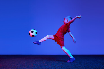 Naklejka premium One sportive child, boy in red uniform playing, training football over dark blue background in neon. Concept of action, sportive lifestyle, team game, health, energy