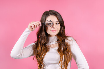 Young beautiful woman holding a magnifying glass near her eye on the pink background. 