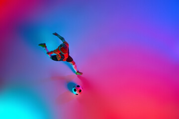 Fototapeta na wymiar Top view of playing little boy during training with football ball in gradient red-blue neon light. Concept of action, sportive lifestyle, team game, health, energy