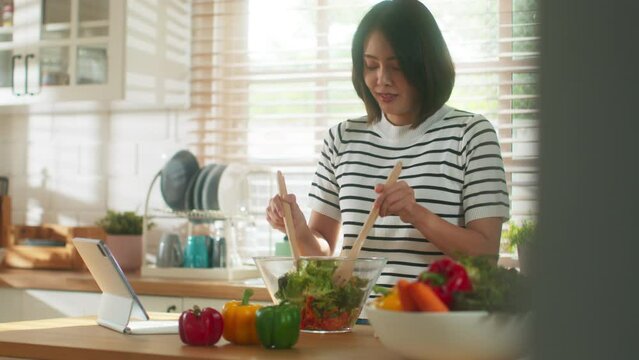 Beautiful young Asian woman hand finger slide tablet screen following healthy vegetarian food recipe in online cooking course. Attractive housewife mixing salad bell pepper, tomato, lettuce together