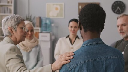 Back view of young Black male patient being supported by female psychology specialist during group therapy session in bright modern office