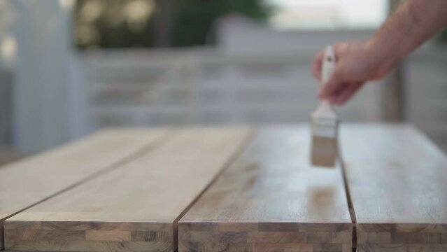 Close-up wooden table with male Caucasian hand applying primer and leaving. Closeup unrecognizable man painting carpentry outdoors