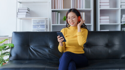 Joyful Young Asian Woman Enjoying Music and Games on Smart Phone at home. Asia female in yellow shirt wearing headphones using mobile phone enjoying listening to music and playing games at home.