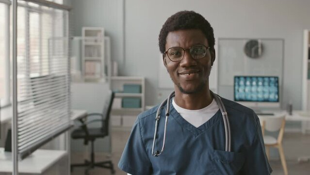 Tilt up portrait of young cheerful Black male physician wearing blue scrubs and stethoscope on his shoulders taking notes on clipboard and posing for camera standing in bright modern doctor office