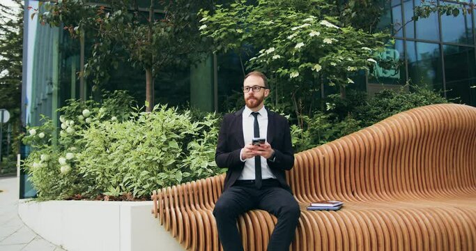 Handsome bearded man using typing mobile phone sitting on bench on city street. Attractive businessman on the phone in an office building. Businessman texing on phone.