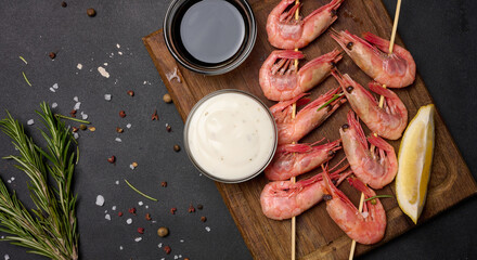 Shrimps strung on wooden sticks, soy sauce and spices on a black table, top view